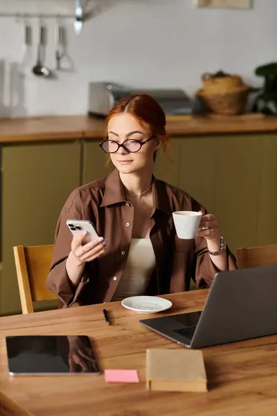 A red haired woman sits at a wooden table with coffee, focused on her smartphone. — Stock Photo