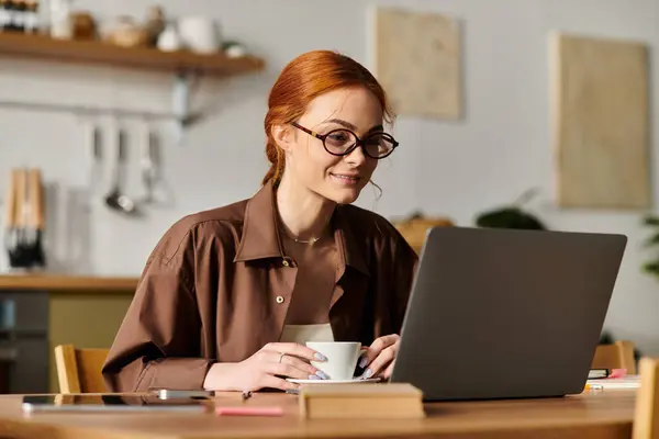 A red haired woman smiles while participating in a digital meeting, sipping coffee at a cafe. — Stock Photo