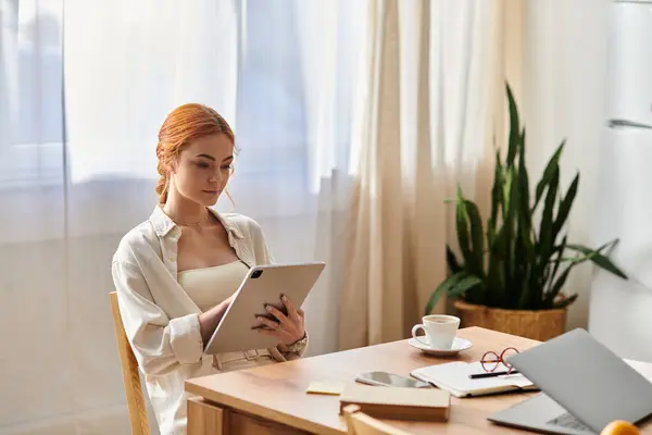 Sunlight fills the room where a red haired woman relaxes with her tablet at a wooden table. — Stock Photo