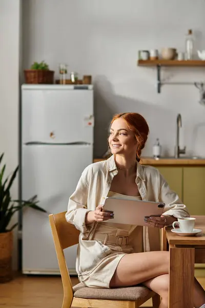 Una donna dai capelli rossi sorseggia un caffè e legge una nota su un accogliente tavolo da cucina.. — Foto stock