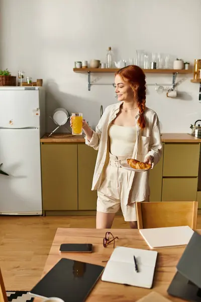 Eine fröhliche rothaarige Frau in einer gemütlichen Küche hält ein Getränk und Gebäck für ihren Tag bereit. — Stockfoto