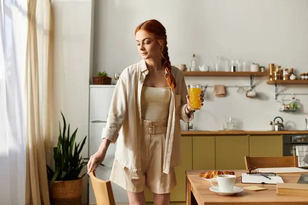 A beautiful woman with red hair stands gracefully in a sunny kitchen, holding refreshing juice. — Stock Photo