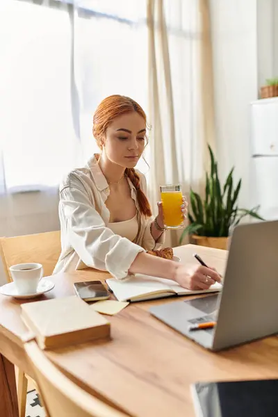 A woman with vibrant red hair writes notes while sipping juice, surrounded by a cozy kitchen. — Stock Photo