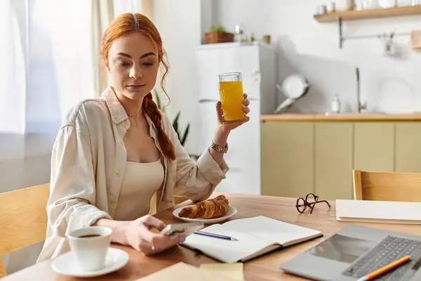 Eine Frau mit leuchtend roten Haaren sitzt bei einem Drink und Croissant in ihrer morgendlichen Routine. — Stockfoto
