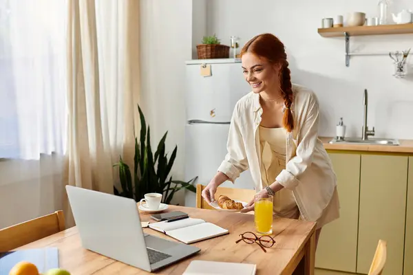 Una donna dai capelli rossi allegra si prepara ad assaporare la sua colazione mentre si impegna con il suo computer portatile. — Foto stock