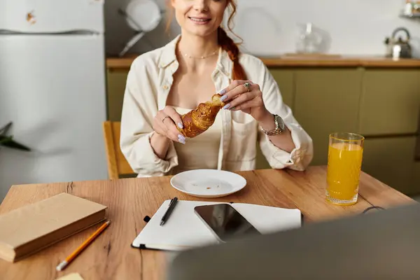 A red haired woman savors a croissant while sipping juice in her modern kitchen at morning light. — Stock Photo