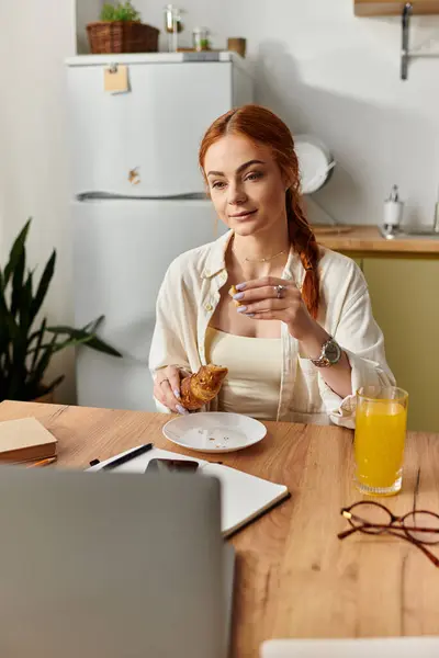 A woman with striking red hair enjoys a pastry during an online meeting in a sunny kitchen. — Stock Photo