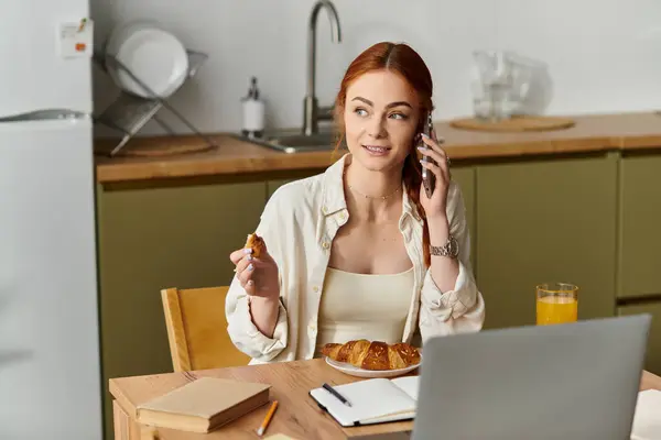 Rothaarige Frau hält ein Croissant in der Hand, während sie an einem Küchentisch ein fröhliches Gespräch führt. — Stockfoto