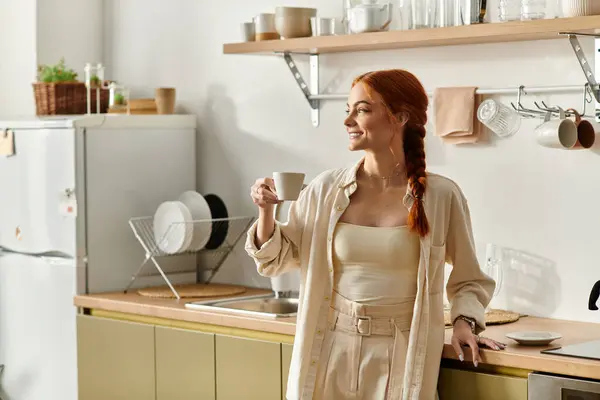 A beautiful red haired woman holds a cup, smiling warmly in her modern kitchen at dawn. — Stock Photo