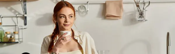 Mujer pelirroja sonríe suavemente mientras sostiene una taza en su elegante cocina, inmersa en el pensamiento. - foto de stock