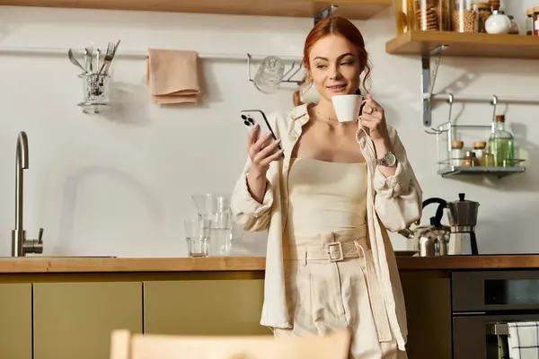 A beautiful red haired woman relaxes in her kitchen, sipping coffee and scrolling on her phone. — Stock Photo