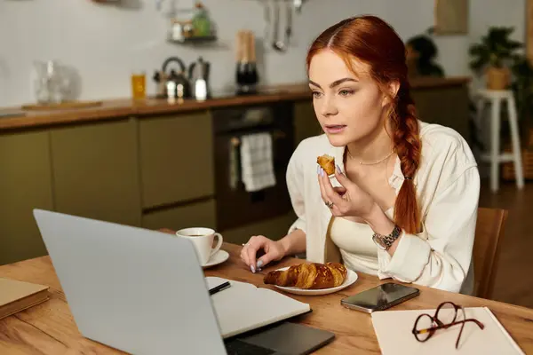 Una donna con i capelli rossi gode di dolci a un tavolo di legno durante l'utilizzo del suo computer portatile. — Stock Photo