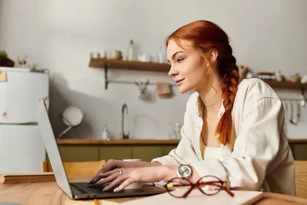 Focused red haired woman types on laptop in a cozy kitchen filled with natural light. — Stock Photo