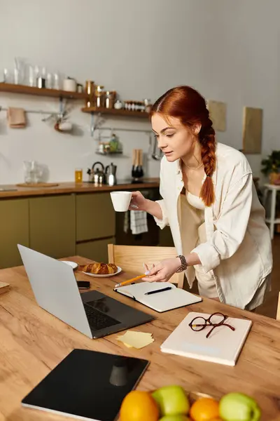 Focused woman with red hair balances a cup while taking notes at her kitchen table with a laptop. — Stock Photo