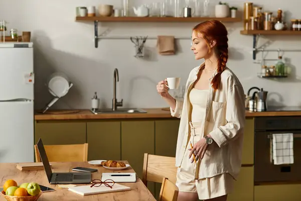 In a modern kitchen, a red haired woman stands holding a cup of coffee, focused on her laptop. — Stock Photo