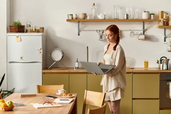 Una donna dai capelli rossi si trova in una cucina moderna, focalizzata sul suo computer portatile mentre assapora il momento. — Foto stock
