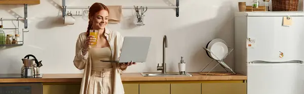 A joyful red haired woman stands in a stylish kitchen, sipping a drink while using her laptop. — Stock Photo