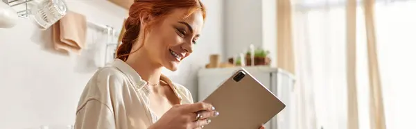 Светлая женщина с рыжими волосами наслаждается планшетом в уютной кухне, наполненной теплом.. — стоковое фото