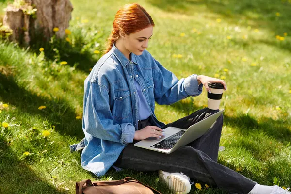 Red haired woman sits on grass, sipping coffee and typing on her laptop in a sunny park. — Stock Photo