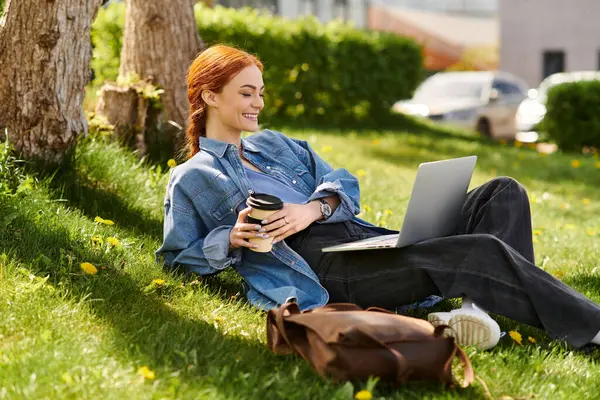 A woman with beautiful red hair relaxes on grass, sipping coffee and typing on her laptop joyfully. — Stock Photo