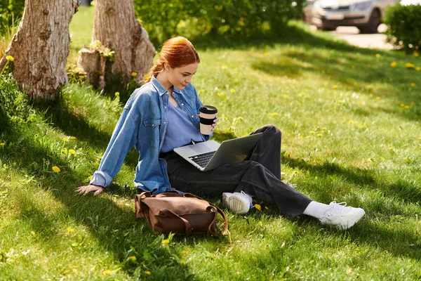 Young woman with striking red hair sits on grass, focused on her laptop while enjoying coffee. — Stock Photo
