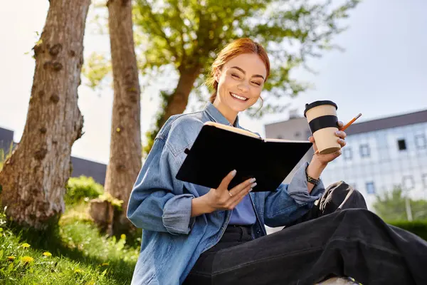A cheerful woman with beautiful red hair sits on the grass, reading a book while sipping coffee. — Stock Photo