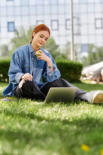 A beautiful woman with red hair casually types on her laptop while savoring an apple in a lush park. — Stock Photo