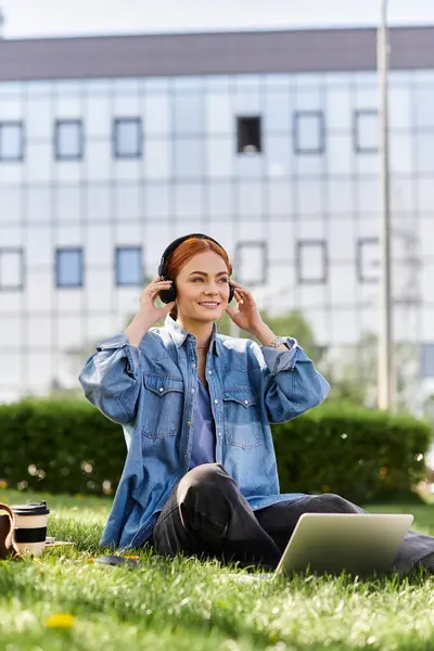 Una donna dai capelli rossi allegra si rilassa sull'erba, impegnata con il suo computer portatile e le cuffie. — Foto stock
