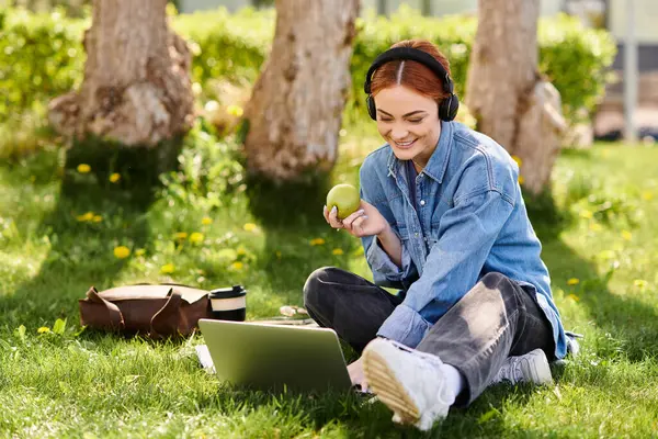 Beautiful woman with red hair sits on grass, engaging with her laptop in a cheerful outdoor setting. — Stock Photo
