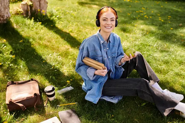 A cheerful red haired woman studies on the grass with headphones, relaxing outdoors. — Stock Photo