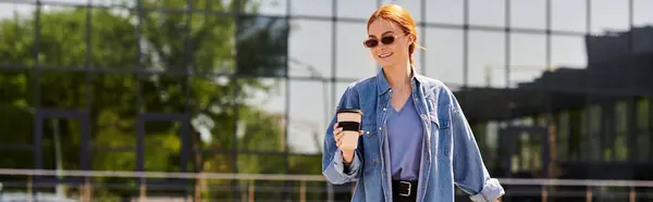 A beautiful red haired woman walks confidently with a coffee cup in hand, smiling in the sunlight. — Stock Photo