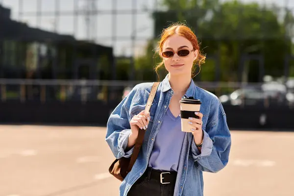 Mujer pelirroja con atuendo casual sonríe mientras sostiene una bebida fuera de una estructura contemporánea. - foto de stock