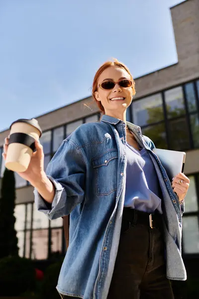 Brightly smiling woman with red hair holds a coffee while standing in front of a stylish building. — Stock Photo