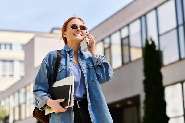 A beautiful woman with red hair smiles while walking and talking on her phone in an urban setting. — Stock Photo