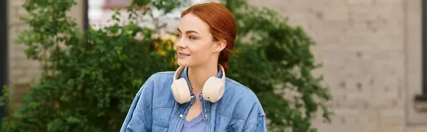A red haired woman strolls happily through a sunny park, wearing headphones. — Stock Photo
