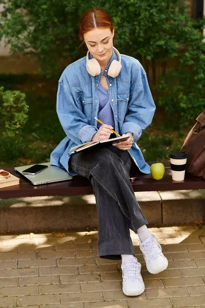 Stylish woman with red hair sits on a bench, sketching in her notebook while savoring coffee. — Stock Photo