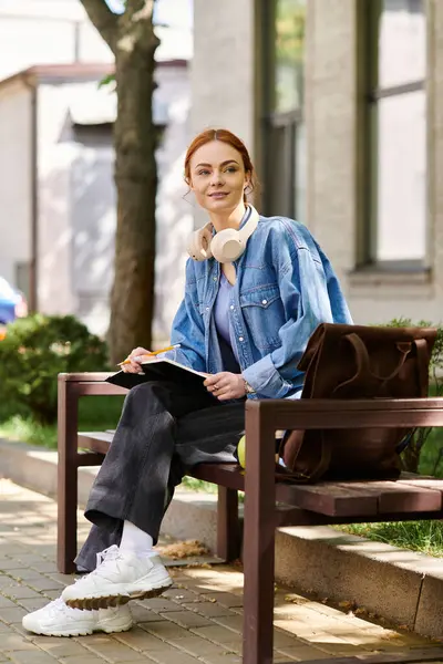 Red haired woman sitting casually on a bench, immersed in writing with a warm smile. — Stock Photo