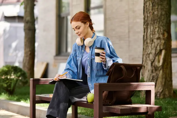 A beautiful red haired woman sits on a bench, sipping coffee and writing in her notebook. — Stock Photo