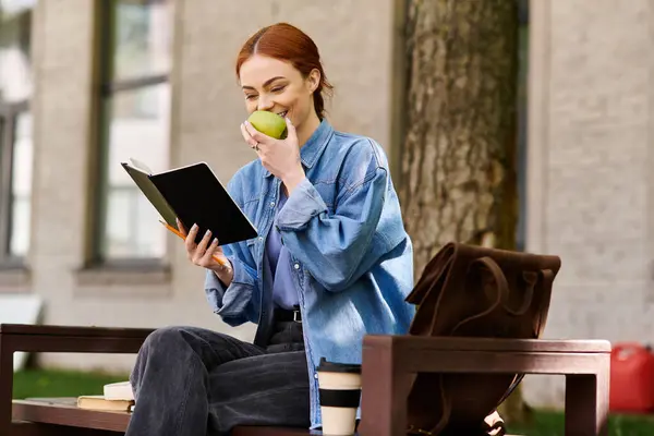 A red haired woman sits on a bench, savoring an apple while reading outdoors. — Stock Photo