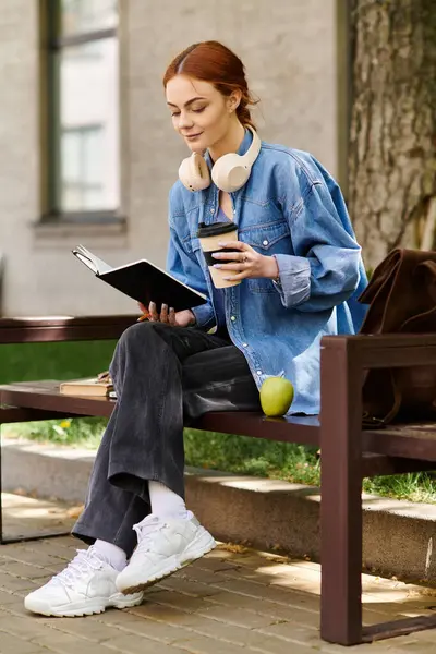 Sitting on a bench, a beautiful red haired woman immerses herself in a book, sipping coffee. — Stock Photo