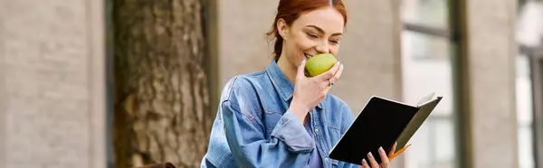 Eine schöne rothaarige Frau lächelt, als sie ein Buch liest, während sie einen grünen Apfel in der Hand hält. — Stockfoto