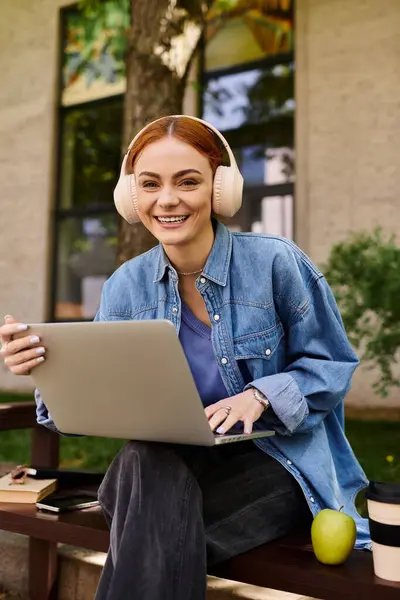 Red haired woman smiles bright with her laptop outdoors on a sunny day, apple beside her. — Stock Photo