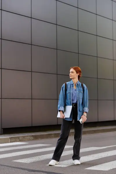 A stylish red haired woman in a denim jacket gazes thoughtfully while crossing the street. — Stock Photo