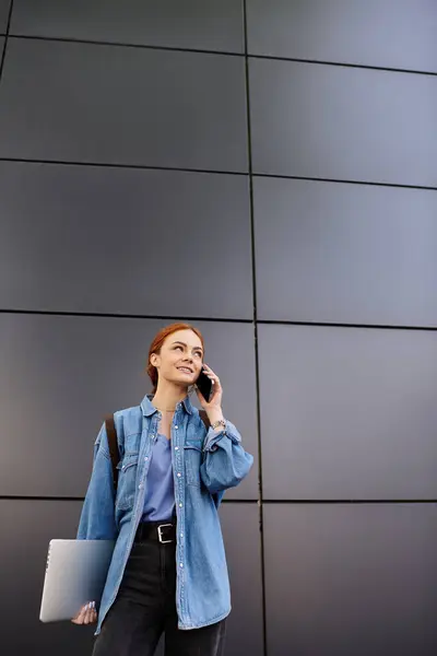 Mulher alegre com cabelo vermelho se envolve em uma conversa telefônica ao ar livre ao lado de um edifício moderno. — Fotografia de Stock