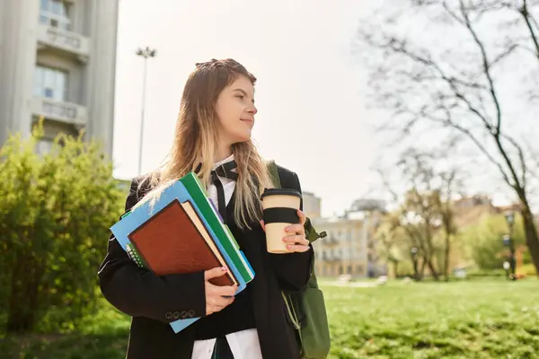 Jolie collégienne en uniforme élégant se tient à l'extérieur, tenant du café et des livres, profiter de la journée. — Photo de stock