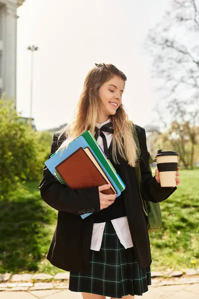 In a vibrant park, a stylish college girl smiles as she carries her books and coffee cup. — Stock Photo