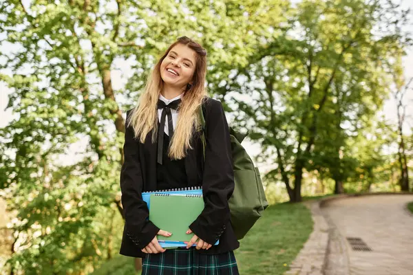 A pretty young adult in a stylish uniform smiles while holding books on a sunlit college path. — Stock Photo