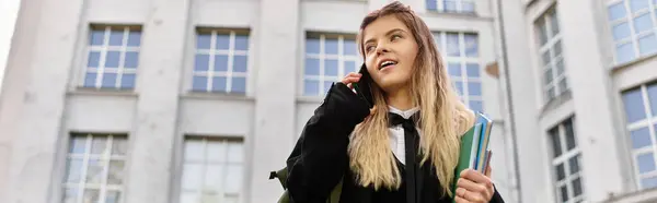 Bright young adult in stylish uniform talks on phone while holding books near college, banner — Stock Photo