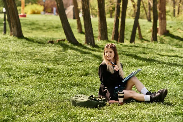 Una bella ragazza del college con un'uniforme elegante si siede sull'erba, abbozzando felicemente nel suo quaderno.. — Foto stock