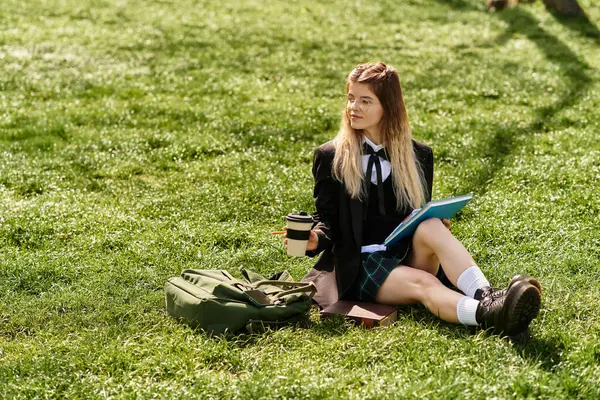 Bella studentessa si rilassa sull'erba, sorseggiando caffè mentre studia i suoi appunti in un'uniforme elegante. — Foto stock